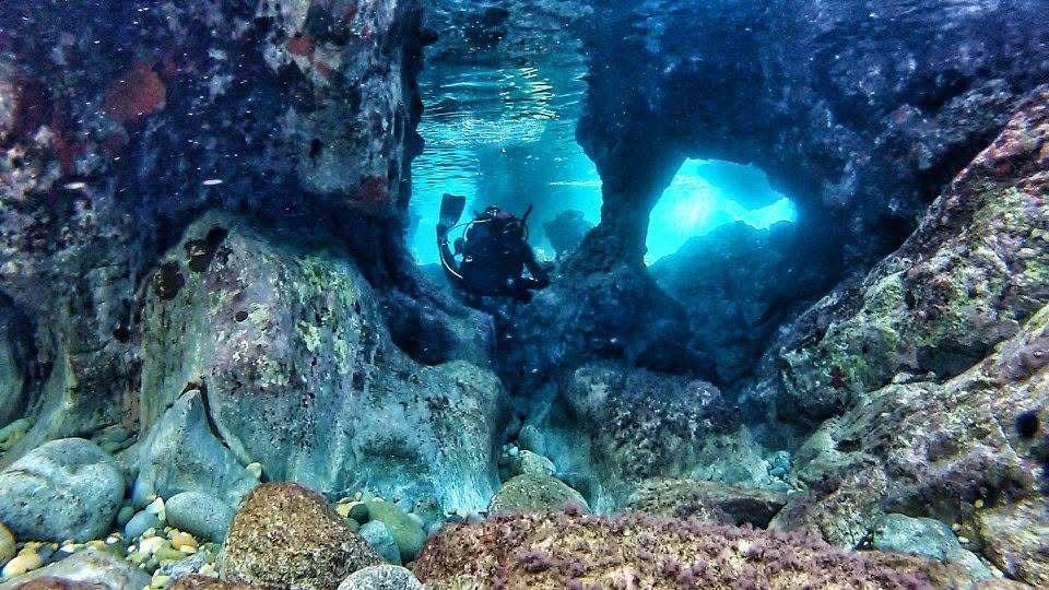 Scuba diver swimming through a natural underwater tunnel formed by large volcanic rocks. Captured during a Dive Greece exploration dive in Halkidiki, Greece. Crystal clear water and light rays create a surreal cave-like view.